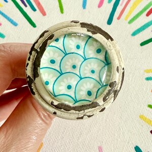 May include: A round cabinet knob with a distressed off-white finish and a central circular inlay featuring a repeating pattern of light blue and white stylized flowers arranged in overlapping scalloped arcs under a smooth glass dome.