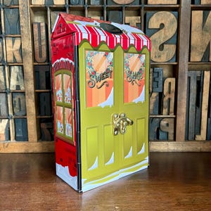 May include: A red and white striped Christmas-themed tin box shaped like a storefront with a gold door and the words "Sweet Shop" on the windows.