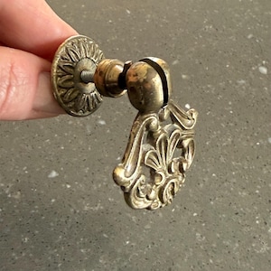 May include: Antique brass drawer pull with a decorative floral design. The pull features a round backplate with a sunburst pattern and a curved handle with intricate detailing. The hardware is made of brass and has a vintage aesthetic.