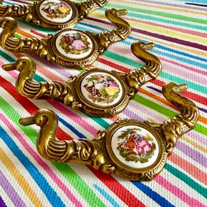 May include: Four brass drawer pulls featuring ornate, curved handles and central circular porcelain insets. Each white porcelain disc displays a colorful, detailed scene of figures in historical attire, set within a decorative metal frame.