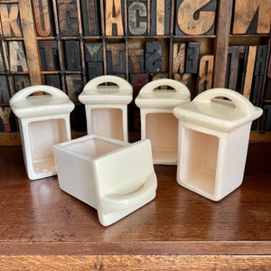 May include: Five off-white ceramic containers with a raised rim and a handle on the top. The containers are arranged in a row on a wooden surface.