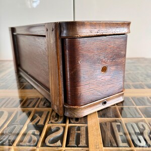 May include: A rectangular dark brown wooden box with a curved front edge and a small circular hole centred on the front panel. The box features a visible wood grain texture and is constructed with vertical side supports and rounded corners.