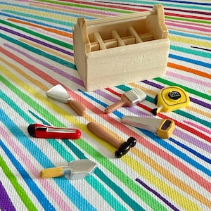 Könnte beinhalten: Eine Miniatur-Werkzeugkiste aus Holz mit verschiedenen Spielzeugwerkzeugen, darunter eine Kelle, ein Pinsel, ein Maßband und eine Säge. Die Werkzeuge sind auf einem farbenfrohen, gestreiften Hintergrund angeordnet.