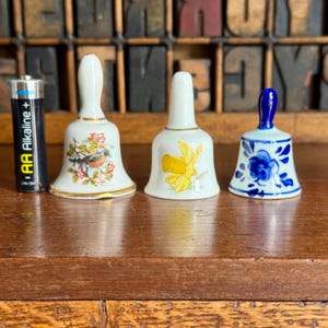 May include: Three decorative ceramic bells with floral and bird designs. The bells are white with gold trim and feature a robin, daffodils, and blue floral patterns. An AA battery is next to the bells for size comparison.