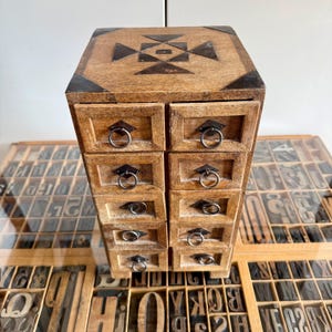 Peut inclure: Une commode haute en bois dotée de dix petits tiroirs disposés en deux colonnes verticales. Chaque tiroir est muni d'une poignée en anneau en métal foncé et d'une plaque en forme de losange. Le dessus présente un motif géométrique en métal.