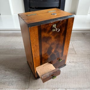May include: A vintage wooden cabinet with a key lock and two small drawers. The cabinet is made of dark wood with lighter wood accents and brass hardware. One drawer is open, revealing a cork insert.