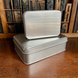 May include: Two silver metal rectangular boxes stacked on top of each other. The top box is slightly smaller than the bottom box.