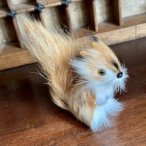 Peut inclure: Un petit jouet en peluche en forme de renard avec une queue touffue, un pelage brun et blanc et des yeux noirs. Le jouet est posé sur une surface en bois.