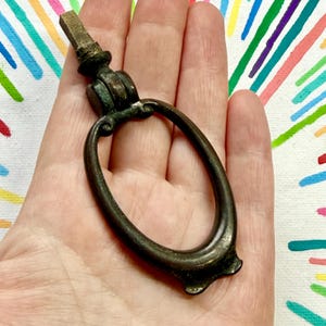 May include: A vintage-style metal drawer pull featuring an oval-shaped drop handle attached to a square-shaped mounting post. The hardware has a dark, weathered patina with visible signs of oxidation and age, showcasing a classic drop-ring design.