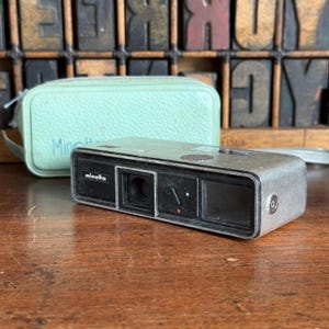 May include: A vintage Minolta camera with a silver body and black accents, next to a light green case. The camera has the brand name "Minolta" printed on the front. The case also has the brand name printed on it.