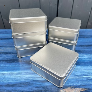 May include: A stack of five silver metal square boxes on a blue wooden surface. The boxes are all the same size and have a smooth, shiny finish.