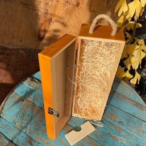 May include: A wooden box with a hinged lid and a handle. The box is open and filled with shredded paper. The box is made of light wood and has a natural finish.