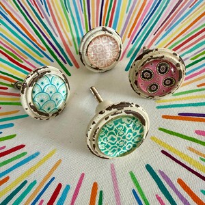 May include: Four round cabinet knobs with distressed white metal edges and glass inserts. Each knob features a unique pattern: one with blue scales, one with pink floral motifs, one with a light pink geometric print, and one with a teal pattern.