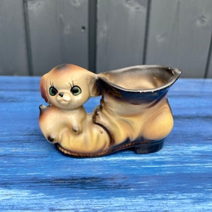 May include: A ceramic planter shaped like a brown boot with a puppy peeking out of the top. The puppy has brown and white fur, big green eyes, and a pink nose.