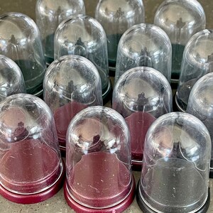 May include: A selection of glass cloches with burgundy and black bases. The clear, dome-shaped covers sit on the bases, creating a display of multiple decorative pieces. The cloches are arranged closely together, showcasing their design.