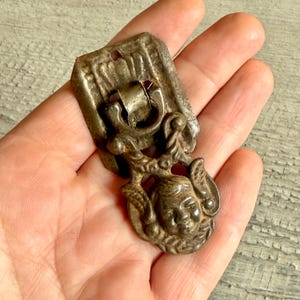 Könnte beinhalten: Antiker silberfarbener Schubladengriff mit dekorativem Design. Der Griff hat eine rechteckige Rückplatte und ein detailliertes Gesicht mit Verzierungen. Das Metall hat ein angelaufenes Aussehen, was auf Alter und Nutzung hindeutet. Ein kleines Loch ist sichtbar.