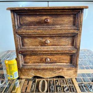 May include: A small, rectangular wooden chest with three horizontal drawers, each featuring a single round wooden knob. The wood has a dark, stained finish with visible grain patterns. A yellow 330ml soda can is placed next to the chest for scale.