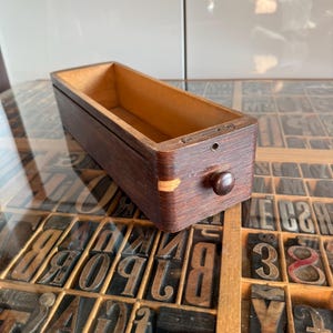 Peut inclure: Un tiroir rectangulaire en bois avec une finition extérieure brun foncé et un intérieur en bois naturel plus clair. La façade présente un petit bouton rond en bois, une vis métallique unique et un petit trou circulaire, avec un éclat visible.