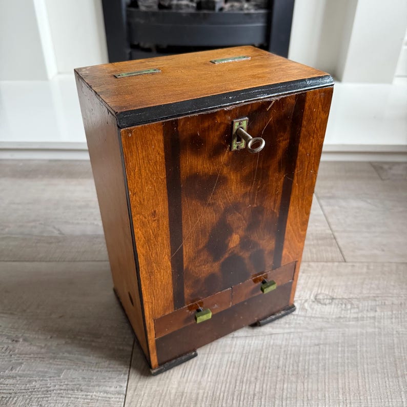 May include: A vintage wooden cabinet with a rectangular shape. The cabinet has a keyhole and a key, two small drawers, and brass hardware. The wood has a warm, reddish-brown tone with darker accents and a black trim.
