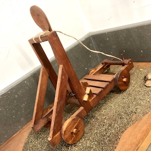May include: A wooden model of a catapult with a rope and a wooden arm. The catapult is on a brown surface with a pile of brown material.
