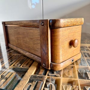 May include: A rectangular wooden box with a small drawer and a round knob. The box is made of light brown wood with darker brown accents and visible wood grain. The box is sitting on a surface with vintage letterpress printing blocks.