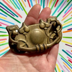 May include: An antique brass drawer pull with intricate detailing. The pull has a curved design with decorative elements and a ring handle. The brass has a warm, aged patina, suggesting vintage charm. The pull is held in a hand against a colourful background.
