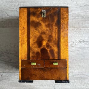 May include: A vintage wooden cabinet with a keyhole and two small drawers. The cabinet is made of wood with a warm, reddish-brown finish and dark brown accents. The drawers have small, rectangular green handles.