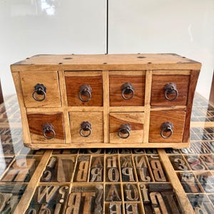 May include: A small wooden chest with eight drawers, each with a decorative metal pull. The wood has a natural finish with varying shades of light and dark brown. The chest sits on a surface with vintage letterpress printing blocks.