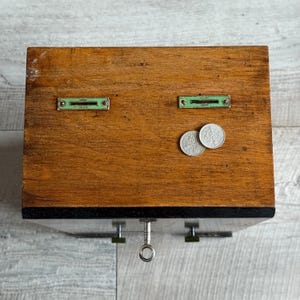 May include: A vintage wooden cash register with two coin slots and a key lock. The register is made of stained wood and has two small metal plates with green accents. Two silver coins rest on the wooden surface.