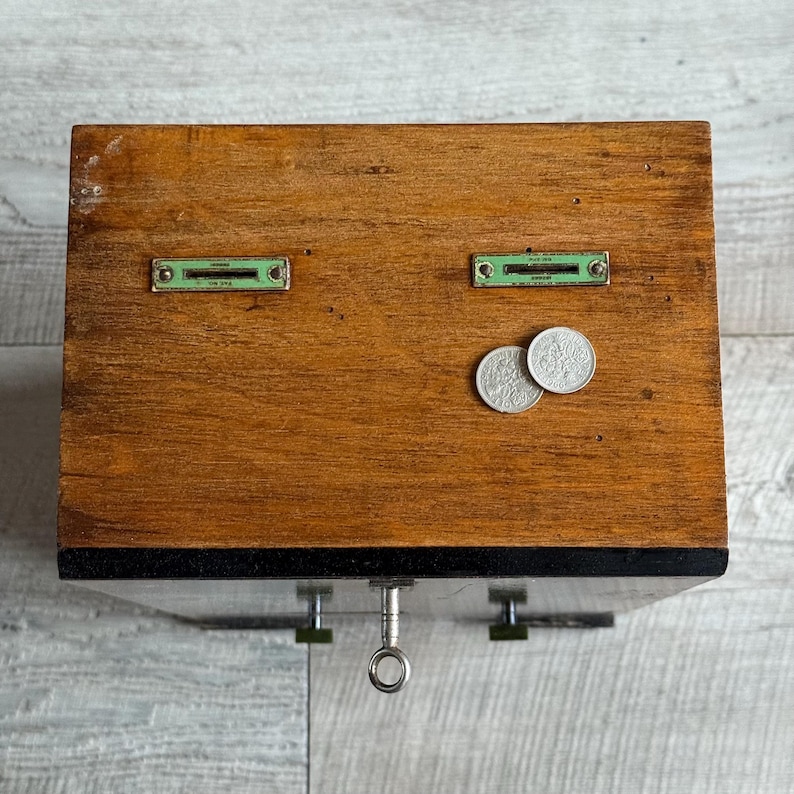 May include: A vintage wooden cash register with two coin slots and a key lock. The register is made of stained wood and has two small metal plates with green accents. Two silver coins rest on the wooden surface.