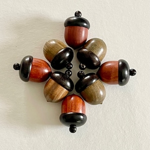 May include: Seven wooden acorn ornaments with black tops. The acorns are arranged in a circle with the tops facing outwards. The acorns are made of different types of wood, including brown, green, and red.