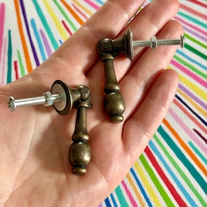 May include: Two antique brass-finished metal drop pulls, each featuring a vertical, rounded handle attached to a circular base plate with a threaded bolt and a silver-coloured metal nut extending from the back for secure installation.