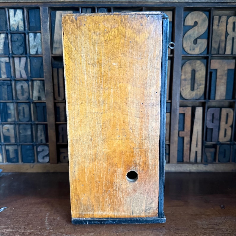 May include: A vintage wooden box with a natural wood grain finish. The box has a rectangular shape with a small circular hole on the front. The edges are outlined in black. The background features a wooden type case with various letters and numbers.