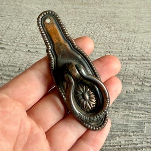 Könnte beinhalten: Antiker Schubladengriff aus Bronze mit floralem Design. Der Griff hat einen Ringgriff und einen detaillierten, perlenbesetzten Rand. Die Hardware ist oval und hat oben ein kleines Loch zur Montage. Das Metall hat ein Vintage-Aussehen.