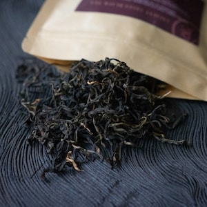 May include: A close-up of a brown paper bag with a label that says "Loose Leaf Black Tea" and a pile of loose leaf black tea on a dark blue surface.
