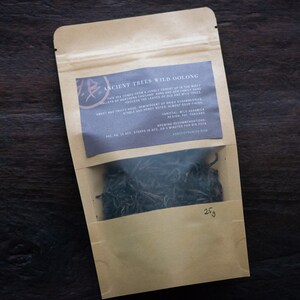 May include: A brown paper pouch with a clear window showing loose green tea leaves. The pouch has a label that reads "Ancient Trees Wild Oolong" and describes the tea as having notes of sweet and fruity, honey, and a hint of wild sesame. The pouch contains 25g of tea.
