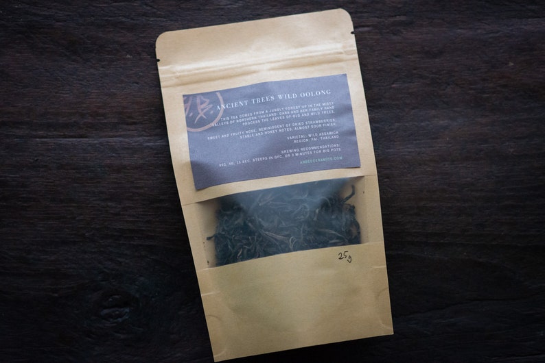 May include: A brown paper pouch with a clear window showing loose green tea leaves. The pouch has a label that reads "Ancient Trees Wild Oolong" and describes the tea as having notes of sweet and fruity, honey, and a hint of wild sesame. The pouch contains 25g of tea.