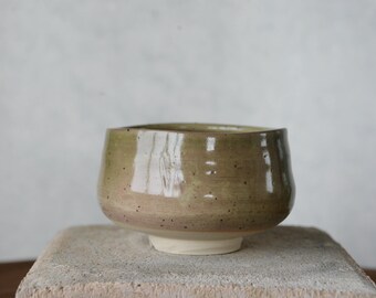 May include: A handmade ceramic bowl with a wide, rounded shape. The bowl has a speckled, earthy green and brown glaze with a white base. The bowl is sitting on a stone surface.