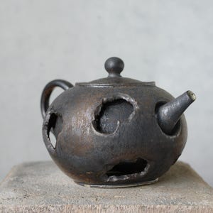 Op de afbeelding: Een handgemaakte, donkergrijze keramische theepot met een ronde body, tuit, handvat en deksel. De theepot heeft unieke, onregelmatige uitgesneden ontwerpen. De theepot staat op een grijs stenen oppervlak.
