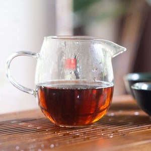 May include: A clear glass teapot with a handle, filled with dark brown tea. The teapot is sitting on a wooden surface with a dark brown teacup in the background.