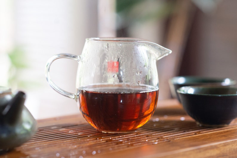 May include: A clear glass teapot with a handle, filled with dark brown tea. The teapot is sitting on a wooden surface with a dark brown teacup in the background.