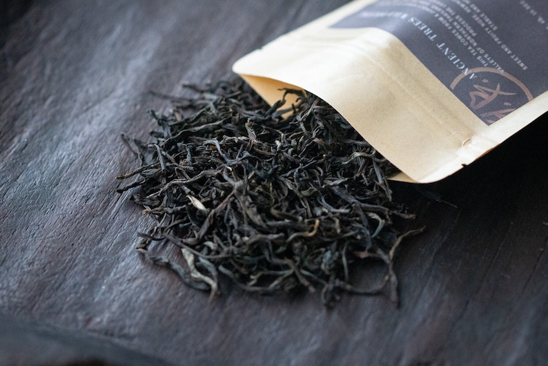 May include: A close-up of loose black tea leaves on a dark wooden surface. A brown paper bag with the text "Ancient Trees" is partially visible in the upper right corner of the image.