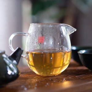 May include: A clear glass teapot with a handle, filled with light brown tea. The teapot is sitting on a wooden surface with a dark green teapot and two dark green teacups.