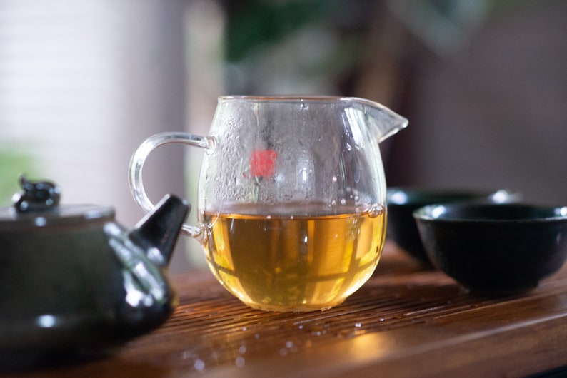 May include: A clear glass teapot with a handle, filled with light brown tea. The teapot is sitting on a wooden surface with a dark green teapot and two dark green teacups.