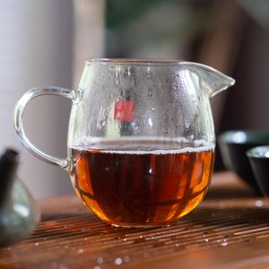 May include: A clear glass teapot with a handle, filled with dark brown tea. The teapot is sitting on a wooden surface with a dark brown teapot and two black teacups.