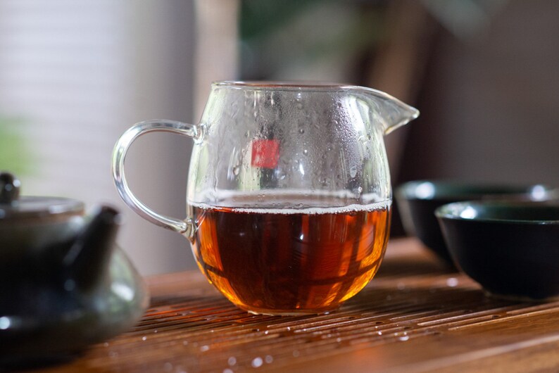 May include: A clear glass teapot with a handle, filled with dark brown tea. The teapot is sitting on a wooden surface with a dark brown teapot and two black teacups.