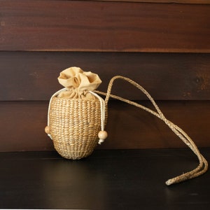 May include: A small, oval-shaped woven bag with a drawstring closure and a long, brown rope handle. The bag is made of natural materials and has a light brown color.
