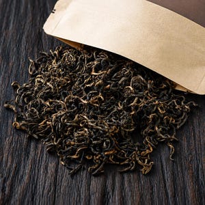 May include: A pile of loose-leaf black tea with dark brown and golden twisted leaves spilling out from an open tan paper pouch. The dried tea leaves are curled and thin, showing a mix of deep brown and lighter golden-brown colour tones.