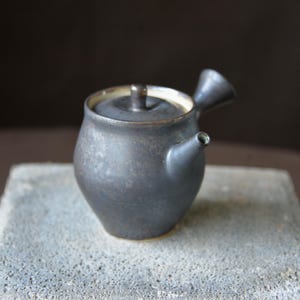 May include: A small, dark gray ceramic teapot with a lid and a spout. The teapot has a rounded body and a handle on the side. The interior is a light color. The teapot is sitting on a gray surface.