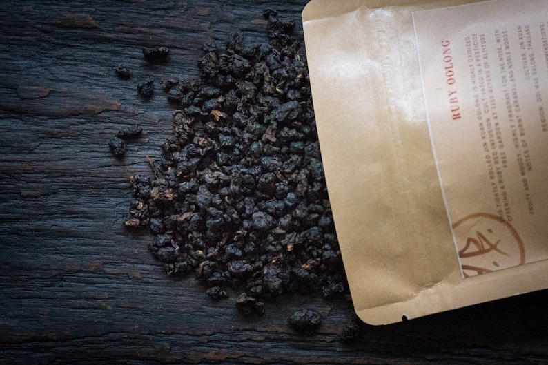 May include: A close-up of a brown paper bag with the text "RUBY OOLONG" printed on it. The bag is filled with loose leaf tea, which is scattered on a dark wooden surface.